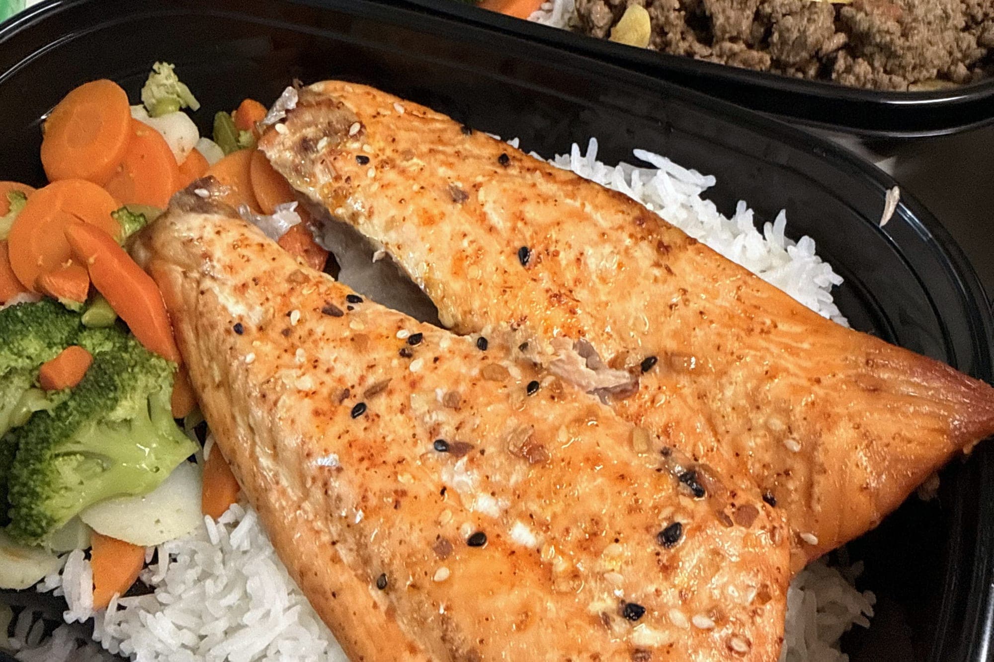 Envase con salmón al horno sobre arroz blanco y vegetales, con un segundo envase detrás